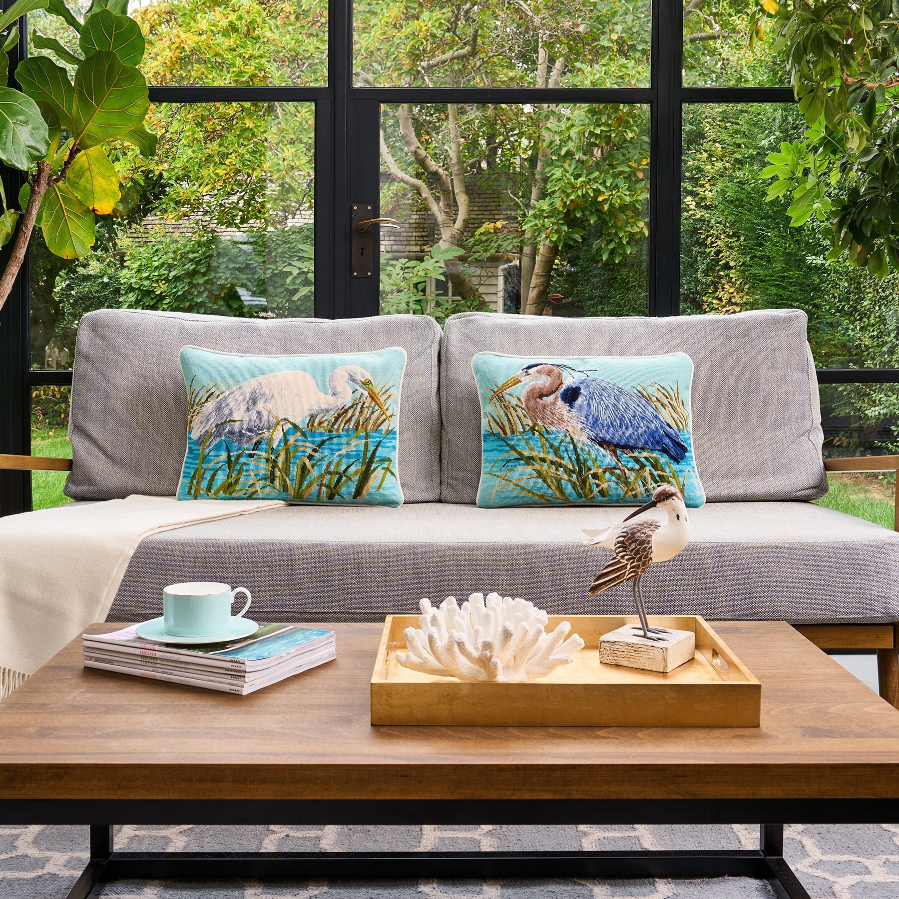 Gray sofa with decorative pillows featuring nature-themed designs, placed in a room with large windows showing greenery outside.