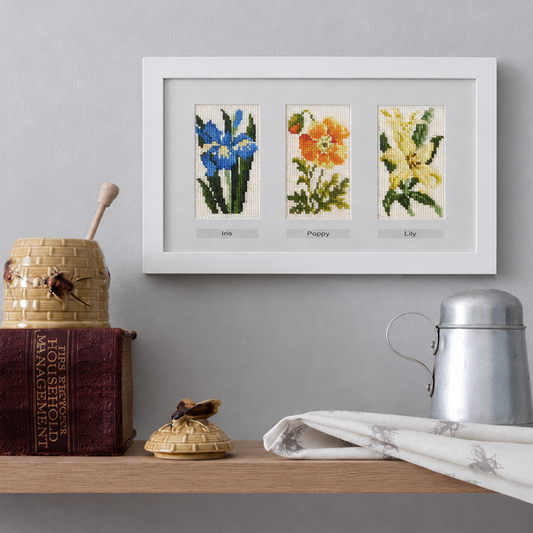 Framed cross-stitch art of flowers in a frame on a shelf with decorative items.