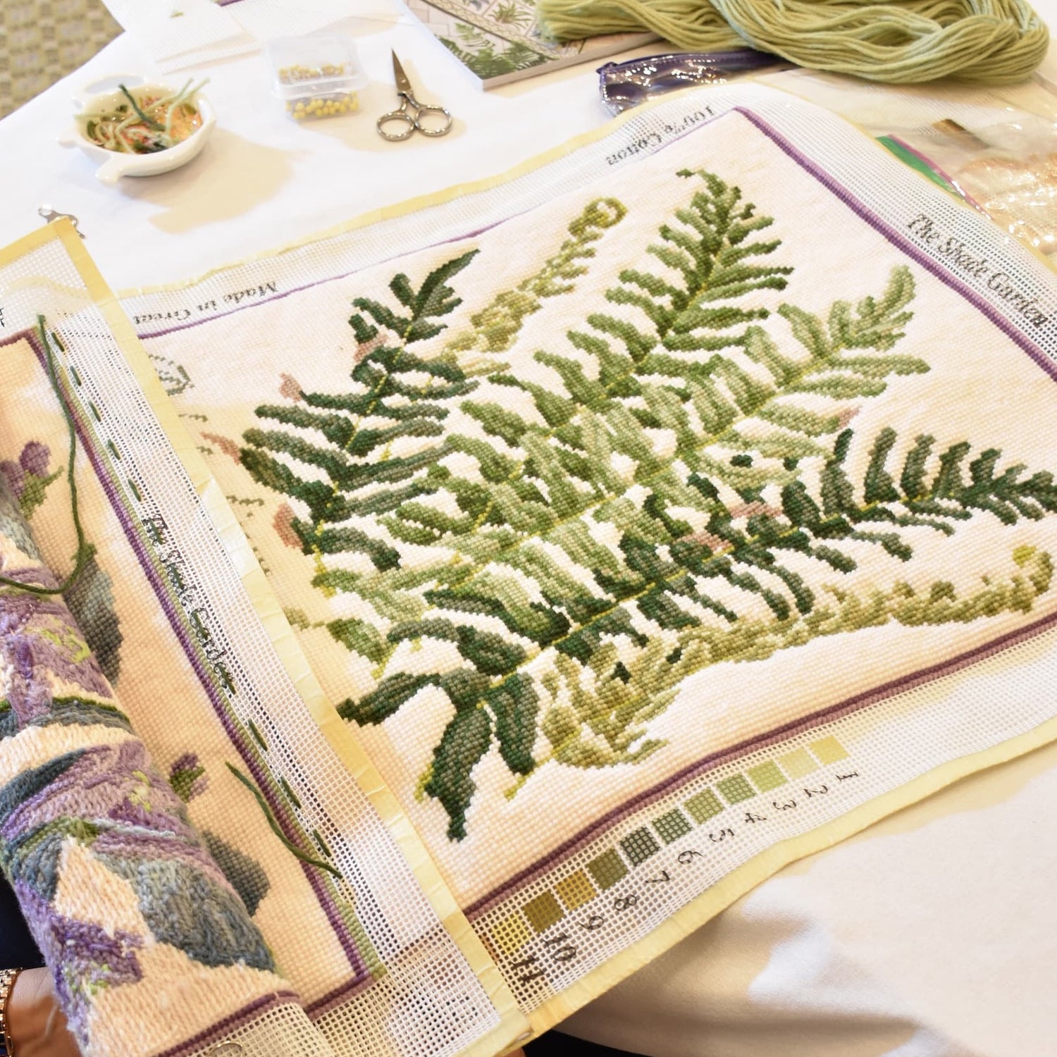A close-up of a needlepoint canvas featuring a green fern design stitched on a cream mesh background, with a color chart strip along the bottom edge and crafting tools nearby on a white table.