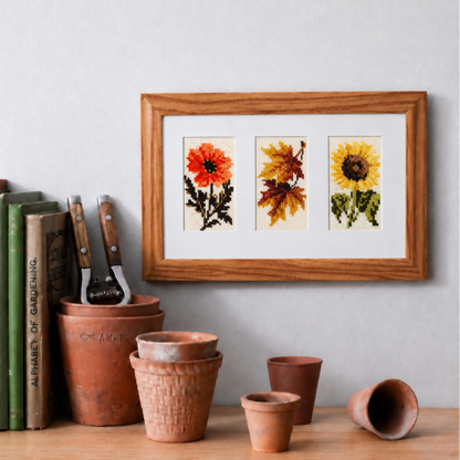 A framed art print featuring three botanical illustrations (a red poppy, autumn leaves, and a yellow sunflower) in a light oak frame, hanging on a light gray wall above a wooden shelf styled with terracotta pots, gardening tools, and vintage books.