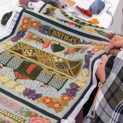 A close-up of a person's hands working on a hand-embroidered tapestry carpet featuring summer motifs and flowers, and the words 'SPRING' and 'SUMMER'.