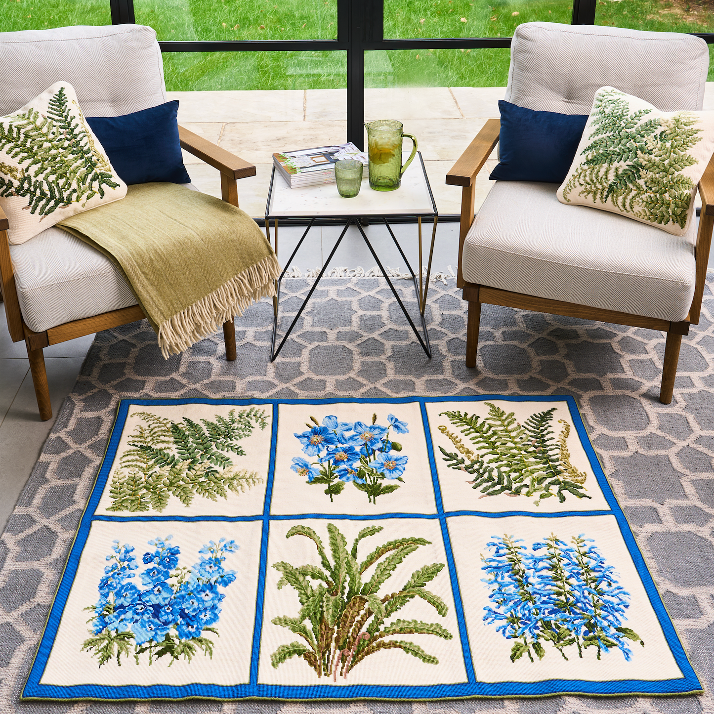 A rectangular area rug with a six-panel botanical design featuring blue flowers and green ferns on a cream background, styled in a modern patio lounge setting between two armchairs.