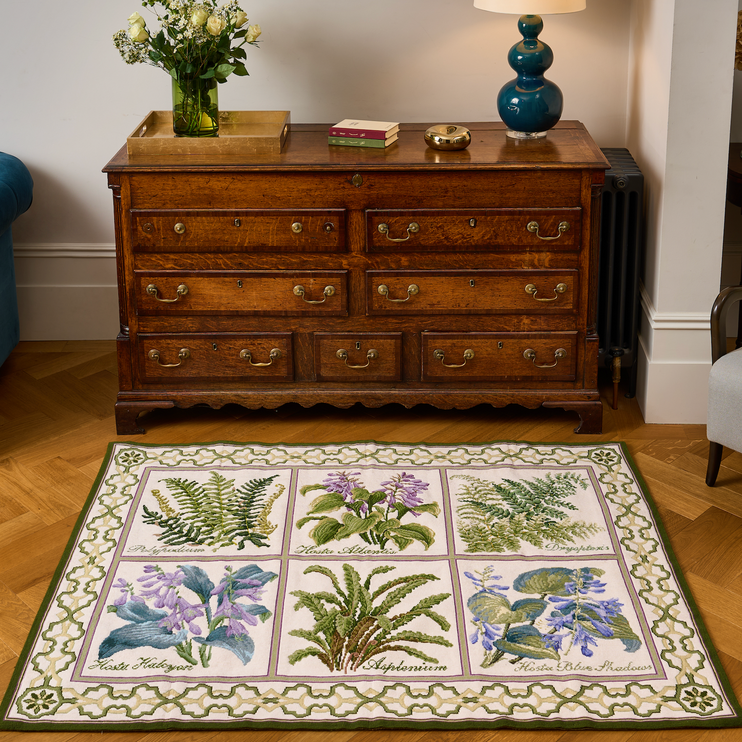 A rectangular area rug with a botanical floral pattern in a 3x3 grid, styled in a living room setting on a wooden parquet floor in front of a dark wood chest of drawers.