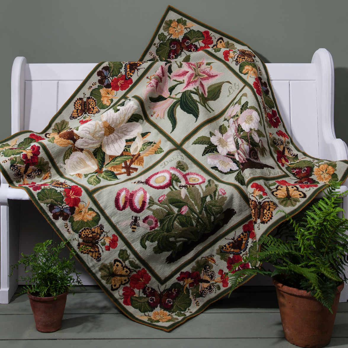 A square floral needlepoint carpet with a pale green background and embroidered flowers, butterflies, and leaves, draped over a white wooden bench with potted ferns on the floor.