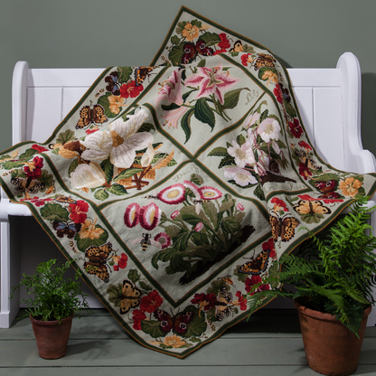 A square floral needlepoint carpet with a pale green background and embroidered flowers, butterflies, and leaves, draped over a white wooden bench with potted ferns on the floor.