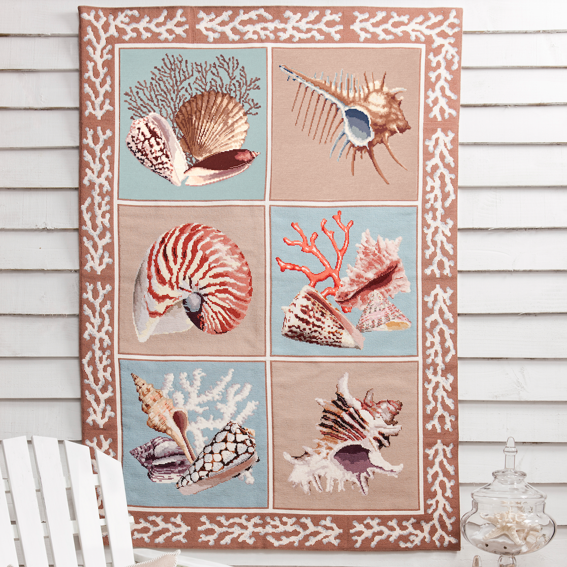 A large rectangular wall tapestry featuring a grid of seashells and coral motifs in beige, pink, blue, and brown tones, hung on a white shiplap wall above a white Adirondack chair with a shell-patterned pillow.