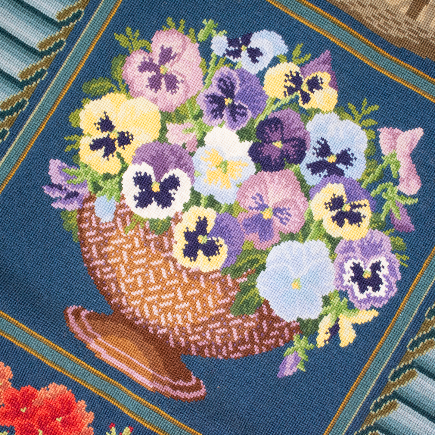 A close-up of a tapestry carpet featuring a basket of pansies in purple, white, yellow, and pink on a dark blue background, with a striped border and small red flowers in the lower left corner.